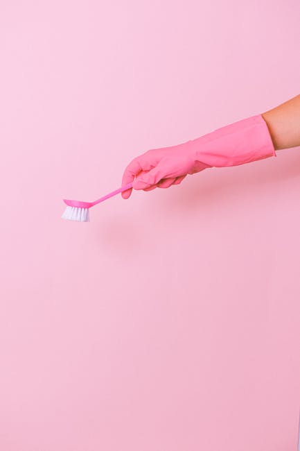 A close-up image of a hand wearing a light pink rubber glove, holding a small pink-handled cleaning brush with white bristles, set against a plain pink background. The scene emphasizes surface cleaning tools used in domestic or commercial sanitisation processes, indicative of thorough cleaning practices carried out by Lambeth Cleaners. The simplicity of the composition highlights attention to hygiene and cleaning procedures applicable to various surfaces such as tiles, countertops, or bathroom fixtures in residential or commercial spaces, reflecting expertise in deep cleaning and surface sanitation.
