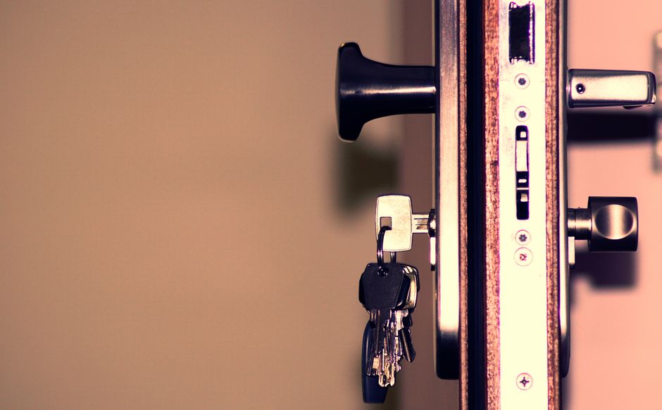A close-up view of a set of metal keys resting on a dark, wet surface, possibly a table or car hood, with reflections visible on the damp surface. The keys include various shapes and sizes, some with